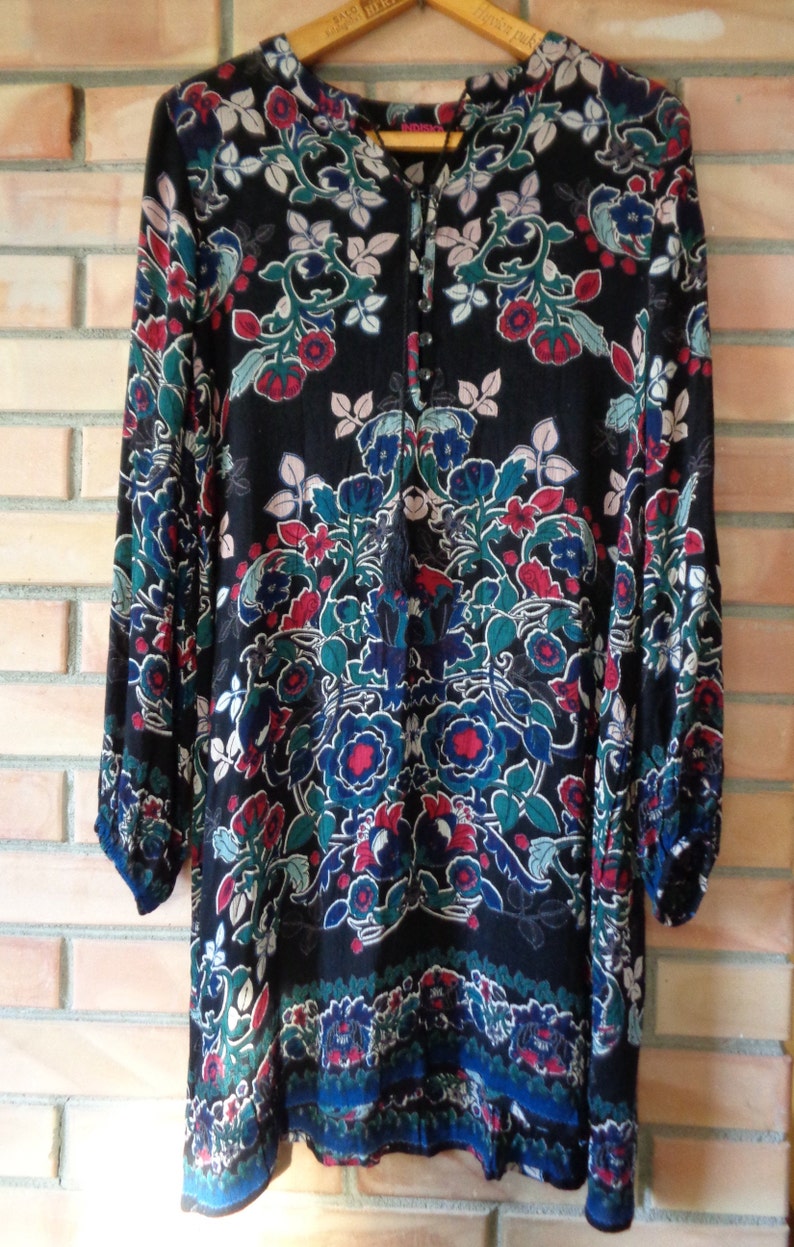 May include: Black tunic dress with a floral pattern in red, blue, teal, and white. The long-sleeved dress has a V-neckline with a tassel tie and button details. The dress is hanging on a wooden hanger.