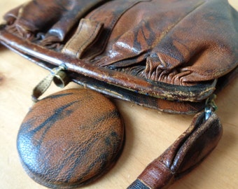 May include: A brown leather purse with a round, brown leather pouch attached. The purse has a flap closure and a metal clasp. The purse is worn and has a vintage look.
