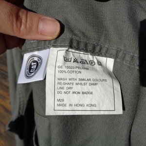 Puede incluir: Una camisa de algod&oacute;n gris con una etiqueta de cuidado blanca. La etiqueta incluye el texto "GE 10022/PELHAM", "100% ALGOD&Oacute;N", "LAVAR CON COLORES SIMILARES", "REMODELAR MIENTRAS EST&Aacute; H&Uacute;MEDO", "SECAR EN LA CUERDA", "NO PLANCHAR LA INSIGNIA", "M29" y "HECHO EN HONG KONG".