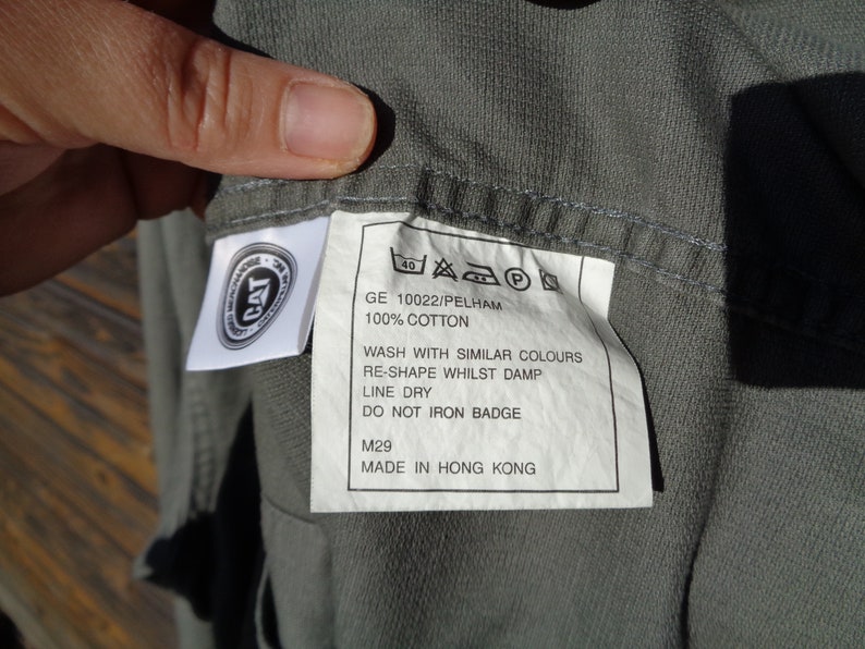 Puede incluir: Una camisa de algod&oacute;n gris con una etiqueta de cuidado blanca. La etiqueta incluye el texto "GE 10022/PELHAM", "100% ALGOD&Oacute;N", "LAVAR CON COLORES SIMILARES", "REMODELAR MIENTRAS EST&Aacute; H&Uacute;MEDO", "SECAR EN LA CUERDA", "NO PLANCHAR LA INSIGNIA", "M29" y "HECHO EN HONG KONG".