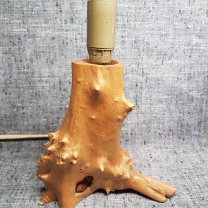 Op de afbeelding: Een handgemaakte houten tafellamp met een natuurlijk, organisch ontwerp. De lamp heeft een lichtbruine, getextureerde basis die lijkt op een boomstronk, met een beige lamphouder aan de bovenkant. De lamp is ontworpen om een standaard gloeilamp te bevatten.