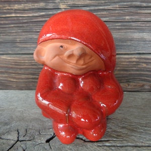 May include: A red ceramic gnome figurine with a smiling face. The gnome is wearing a red hat and coat.