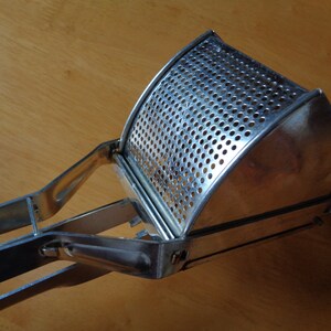 May include: A stainless steel potato ricer with a perforated bowl and long handles. The ricer is designed for mashing potatoes and other soft foods. The kitchen utensil is made of shiny metal and sits on a wooden surface.