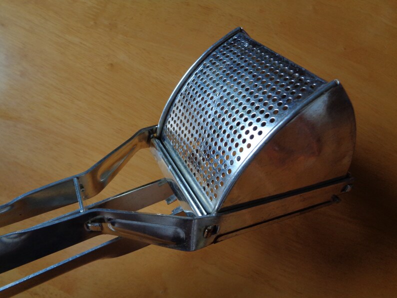 May include: A stainless steel potato ricer with a perforated bowl and long handles. The ricer is designed for mashing potatoes and other soft foods. The kitchen utensil is made of shiny metal and sits on a wooden surface.