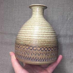 May include: A ceramic vase with a light brown and white speckled glaze. The vase has a wide, rounded body and a narrow neck. The bottom of the vase is decorated with a brown and white geometric pattern.
