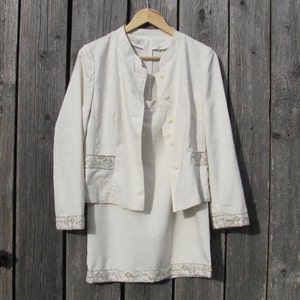 May include: A white linen suit with a jacket and skirt. The jacket has a mandarin collar and a button closure. The skirt has a straight silhouette and a decorative trim around the hem.