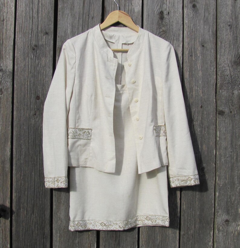 May include: A white linen suit with a jacket and skirt. The jacket has a mandarin collar and a button closure. The skirt has a straight silhouette and a decorative trim around the hem.