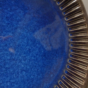 May include: A large, round, blue ceramic plate with a dark brown, textured rim. The plate has a slightly speckled surface.