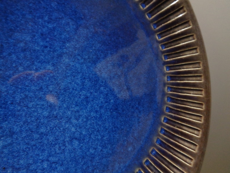 May include: A large, round, blue ceramic plate with a dark brown, textured rim. The plate has a slightly speckled surface.