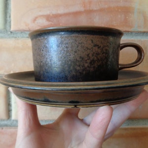 Peut inclure: Une tasse à thé et une soucoupe en céramique brun et noir. La tasse a un glaçage moucheté et une poignée arrondie. La soucoupe a un bord légèrement surélevé.