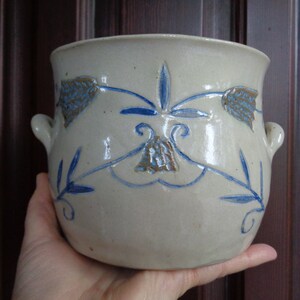 Può includere: Un vaso di ceramica beige con disegni floreali blu e due piccole maniglie. Il vaso ha un'ampia apertura e una superficie leggermente strutturata. Il design blu include foglie e fiori a forma di campana.