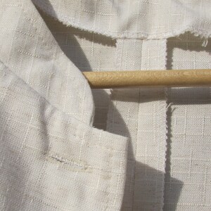 May include: A close-up of a white linen fabric with a seam down the center. The fabric is folded over and has a wooden hanger on top.