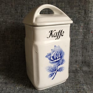 Op de afbeelding: Witte keramische koffiepot met deksel en handvat. Het woord "Kaffe" is in zwart schrift gedrukt boven een blauwe rozenillustratie. De pot heeft een rechthoekige vorm.
