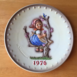 以下が含まれることがあります： 白いセラミック製のプレートに、青いドレスと赤いボンネットを着た女の子が、ピンクの花が咲いた木の枝に座っているレリーフ画像が描かれています。彼女の頭上にある枝には青い鳥がとまっています。プレートは、白い星がついた茶色の縁取りがされており、赤い文字で1976年と記されています。