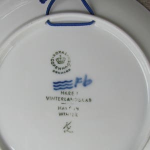 May include: A white ceramic plate with a blue and green Royal Copenhagen logo, a blue wave design, and the text "Hare i Vinterlandskab - Half in Winter".