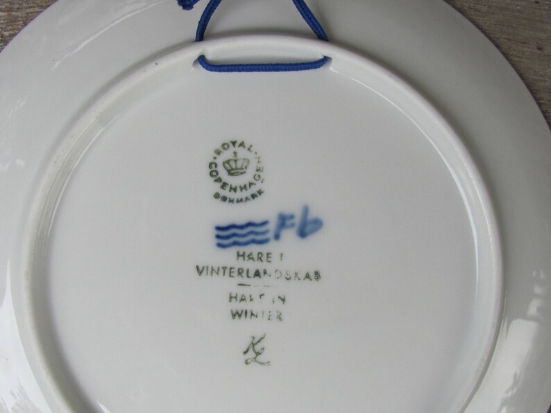 May include: A white ceramic plate with a blue and green Royal Copenhagen logo, a blue wave design, and the text "Hare i Vinterlandskab - Half in Winter".