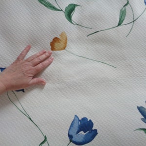 May include: White fabric with a quilted texture and a floral pattern. The flowers are blue and yellow with green leaves.