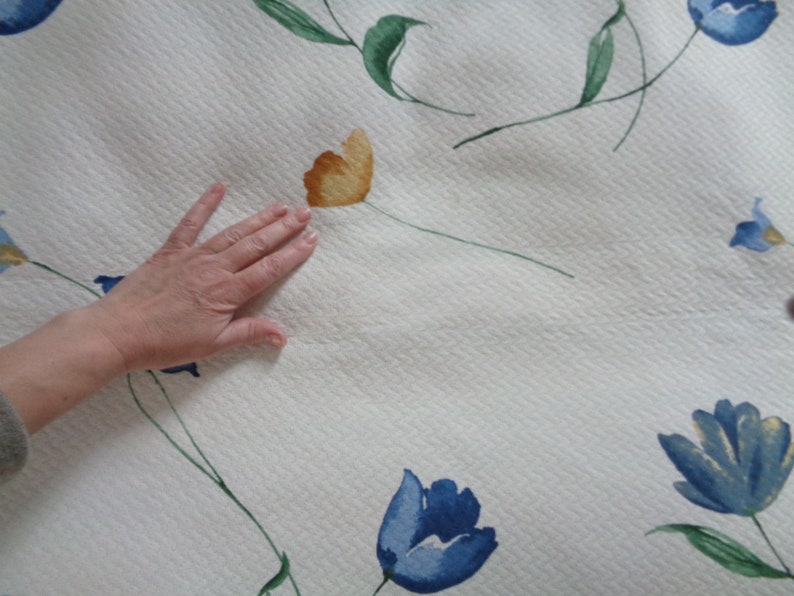 May include: White fabric with a quilted texture and a floral pattern. The flowers are blue and yellow with green leaves.