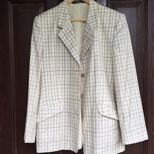 May include: A cream-colored blazer with a plaid pattern in light brown and blue. The blazer has a notched collar, two front pockets, and a single-button closure. It is hanging on a wooden hanger against a dark brown door.