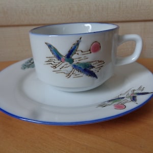 May include: White teacup and saucer set with a blue rim. The cup and saucer feature a bird design in teal, blue, and pink. The cup has a handle and sits on the matching saucer. A charming addition to any tea set collection.