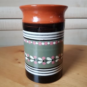 May include: A ceramic vase with a brown, green, white and pink geometric pattern. The vase has a wide mouth and a cylindrical shape.