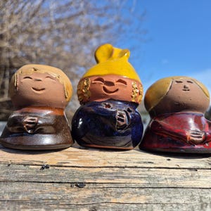 以下が含まれることがあります： 笑顔の顔をした3つのセラミックフィギュア。フィギュアは小さな人の形で、茶色、黄色、赤色の服を着ています。背景は青い空のある木製の表面です。