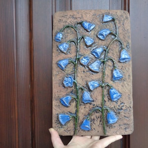 K&ouml;nnte beinhalten: Eine Keramik-Wanddekoration mit einem Reliefdesign aus blauen Glockenblumen auf braunem Hintergrund.