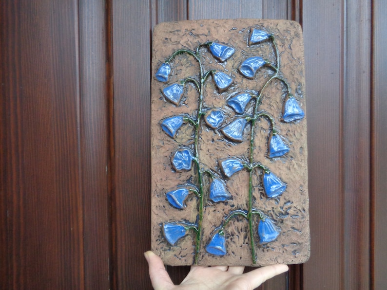 K&ouml;nnte beinhalten: Eine Keramik-Wanddekoration mit einem Reliefdesign aus blauen Glockenblumen auf braunem Hintergrund.