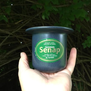 May include: A dark blue ceramic jar of mustard, labeled "Apotekets Senap" in green and white. The jar is held in a hand against a backdrop of green foliage and branches. The label includes text about ingredients and weight.