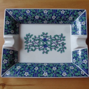Op de afbeelding: Een witte asbak van keramiek met een blauw en groen bloemenmotief. De asbak heeft twee inkepingen aan de zijkanten voor sigaretten.