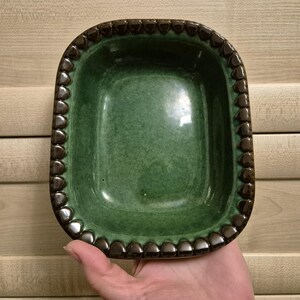 May include: A green ceramic dish with a rectangular shape and rounded corners. The rim is decorated with a series of brown, heart-shaped accents. The dish has a glossy finish and is held in a hand.