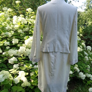 May include: A white linen suit with a skirt and jacket. The jacket has a collar and long sleeves. The skirt is knee-length and has a lace trim. The suit is on a mannequin against a background of white flowers.