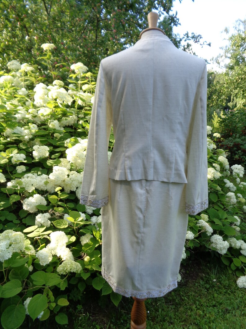 May include: A white linen suit with a skirt and jacket. The jacket has a collar and long sleeves. The skirt is knee-length and has a lace trim. The suit is on a mannequin against a background of white flowers.