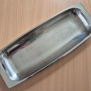 Può includere: Un vassoio rettangolare in acciaio inossidabile con angoli arrotondati. Il vassoio ha una superficie lucida e riflettente ed è vuoto. Il vassoio è progettato per il servizio o l'esposizione di cibo.