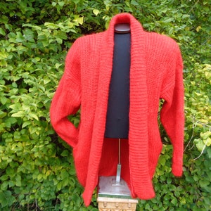 May include: A red knitted cardigan with a shawl collar. The cardigan is made of a soft, fuzzy yarn and has a relaxed fit.