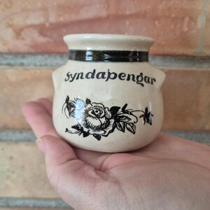 May include: A small, cream-colored ceramic jar with a dark band around the rim and the word "syndapengar" in black script. A black rose and leaf design is below the text. The jar has small handles on each side.