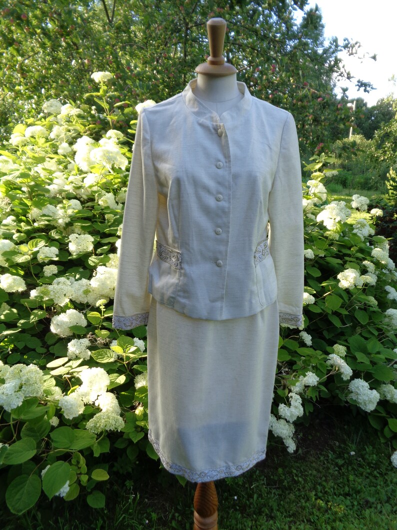 May include: A white linen suit with a skirt and jacket. The jacket has a mandarin collar and buttons down the front. The skirt has a lace trim at the hem.