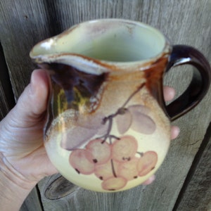 May include: A brown ceramic pitcher with a yellow and brown floral design. The pitcher has a handle and a rounded body. The floral design features a cluster of light pink berries.