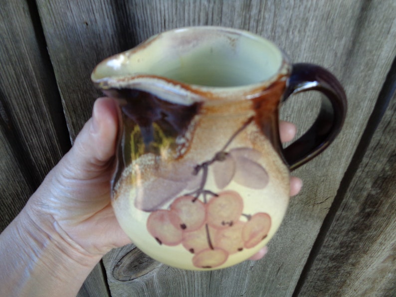 May include: A brown ceramic pitcher with a yellow and brown floral design. The pitcher has a handle and a rounded body. The floral design features a cluster of light pink berries.
