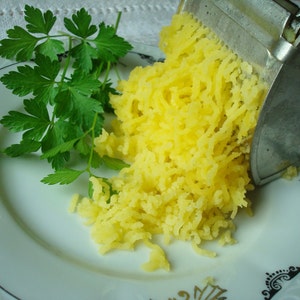 May include: A plate with a sprig of fresh green parsley and a pile of shredded yellow potatoes. A metal potato ricer is positioned over the potatoes, suggesting food preparation. The plate has a decorative rim.