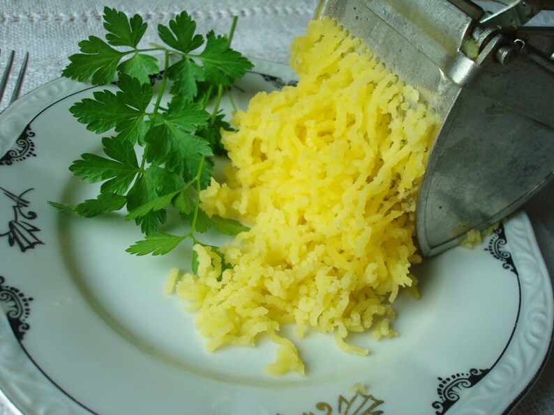 May include: A plate with a sprig of fresh green parsley and a pile of shredded yellow potatoes. A metal potato ricer is positioned over the potatoes, suggesting food preparation. The plate has a decorative rim.