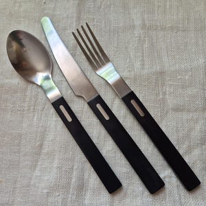 Op de afbeelding: Een roestvrijstalen bestekset bestaande uit een lepel, mes en vork. Elk stuk heeft een zilveren lemmet of tanden en een zwarte handgreep met een rechthoekig zilveren detail. Het bestek ligt op een lichtgekleurde stof.