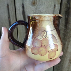 May include: A small, brown and yellow ceramic pitcher with a handle. The pitcher has a painted design of a branch of berries.