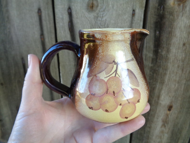 May include: A small, brown and yellow ceramic pitcher with a handle. The pitcher has a painted design of a branch of berries.