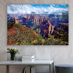 May include: A panoramic photograph of the Grand Canyon, featuring layered rock formations and a cloudy sky. The image is displayed on a canvas and hung on a white wall in a modern living room setting.