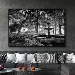 May include: A black and white photograph of a fountain in a park with trees and streetlights. The fountain is in the center of the image and is surrounded by trees and bushes. The streetlights are on either side of the fountain and are casting a soft glow on the scene.