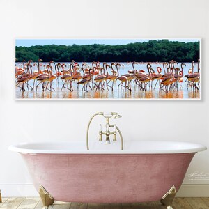 Puede incluir: Una fotograf&iacute;a panor&aacute;mica de una bandada de flamencos rosas de pie en aguas poco profundas. Los flamencos est&aacute;n en fila, mirando hacia la c&aacute;mara. El agua es de color azul claro y el fondo es un bosque verde exuberante. La fotograf&iacute;a est&aacute; enmarcada en un marco blanco y est&aacute; colgada en una pared blanca sobre una ba&ntilde;era rosa.