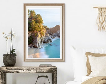 May include: A framed photograph of a waterfall cascading down a rocky cliffside into a turquoise ocean. The photo is hanging on a white wall above a wooden table with a vase of white flowers and a cactus plant.