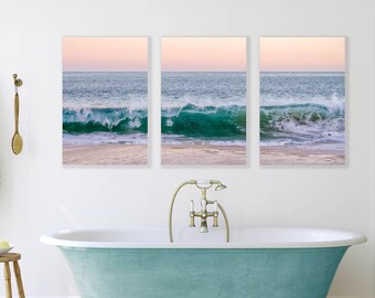 May include: Three framed photographs of a beach scene with a turquoise wave crashing on the shore. The sky is a soft pink and the sand is white. The photos are hanging on a white wall above a turquoise bathtub.