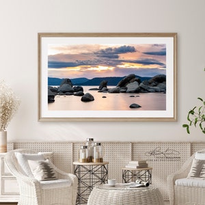 May include: A framed photograph of a sunset over a lake with large rocks in the foreground. The sky is a mix of pink, orange, and purple with clouds. The water is calm and reflects the colors of the sky.